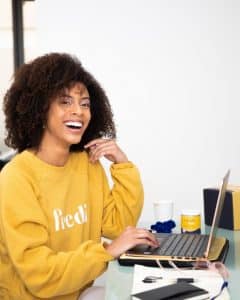 Woman in yellow sweatshirt working on laptop laughing